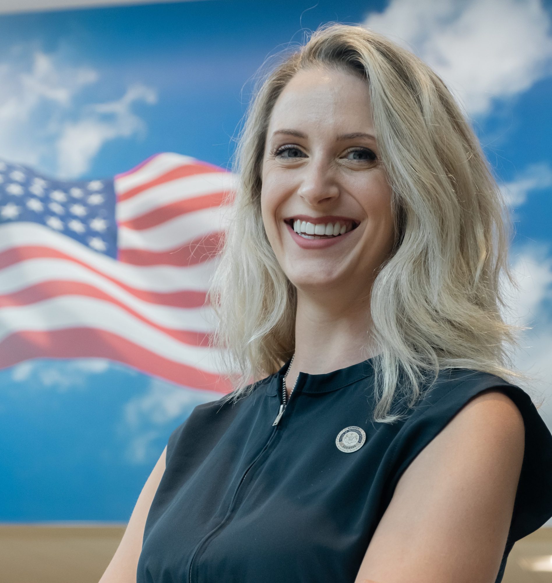 Blonde woman smiling - a mural featuring the flag of the United States is in the background.