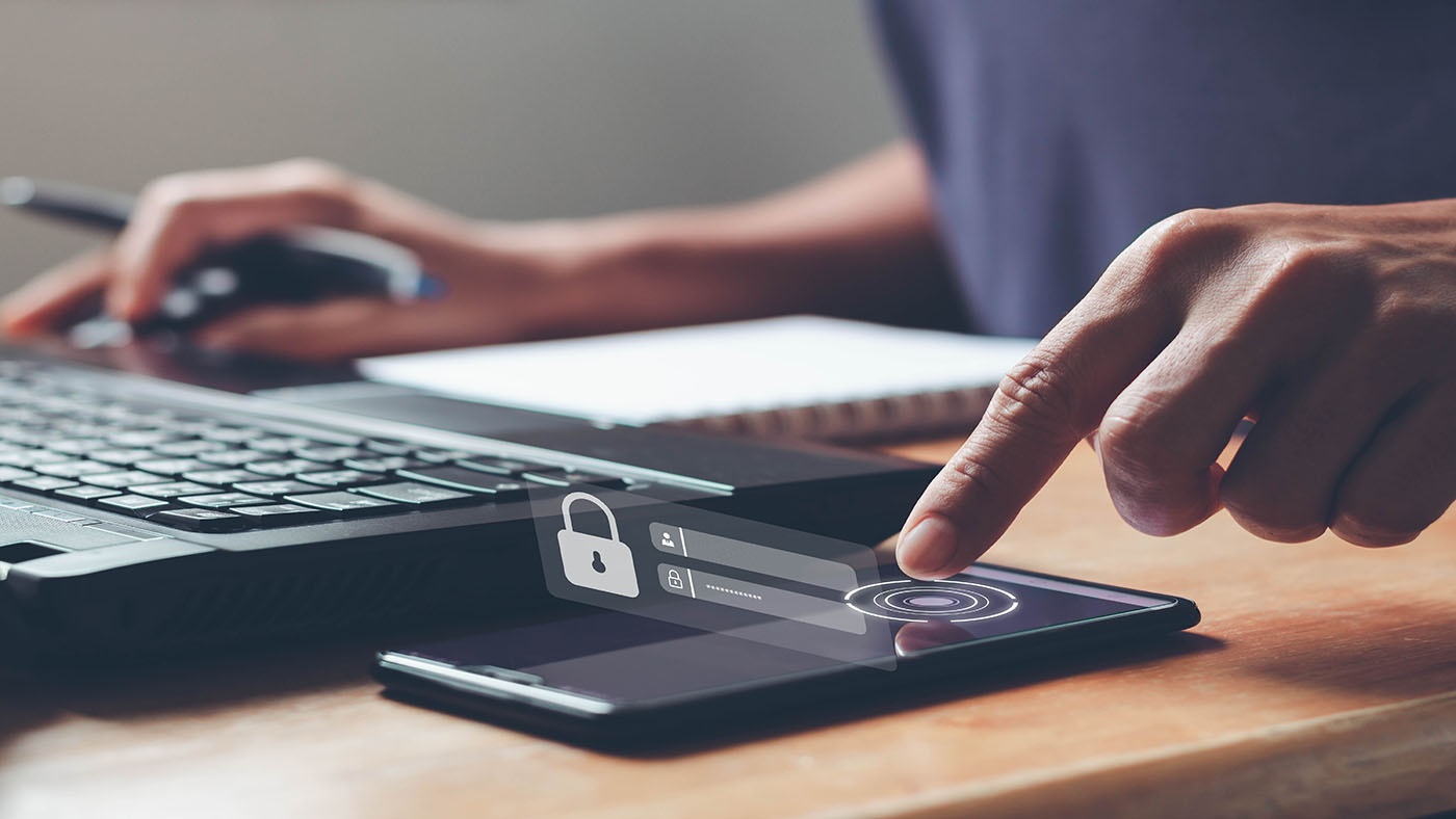 User with both a laptop, paper notebook and mobile device with one hand on the laptop mouse and the other finger selecting the unlock icon on the mobile device.