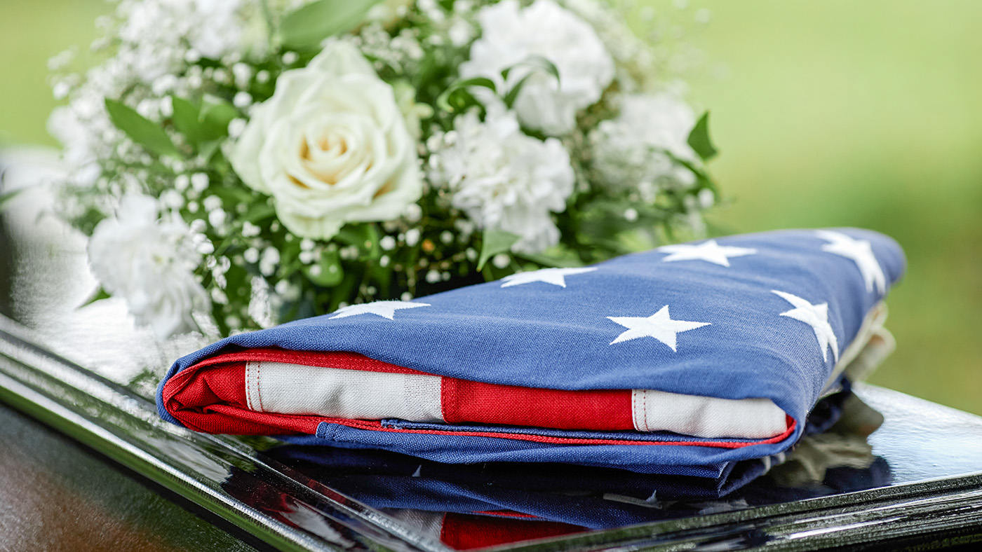Closeup of folded American flag on coffin with flowers at outdoor funeral ceremony.