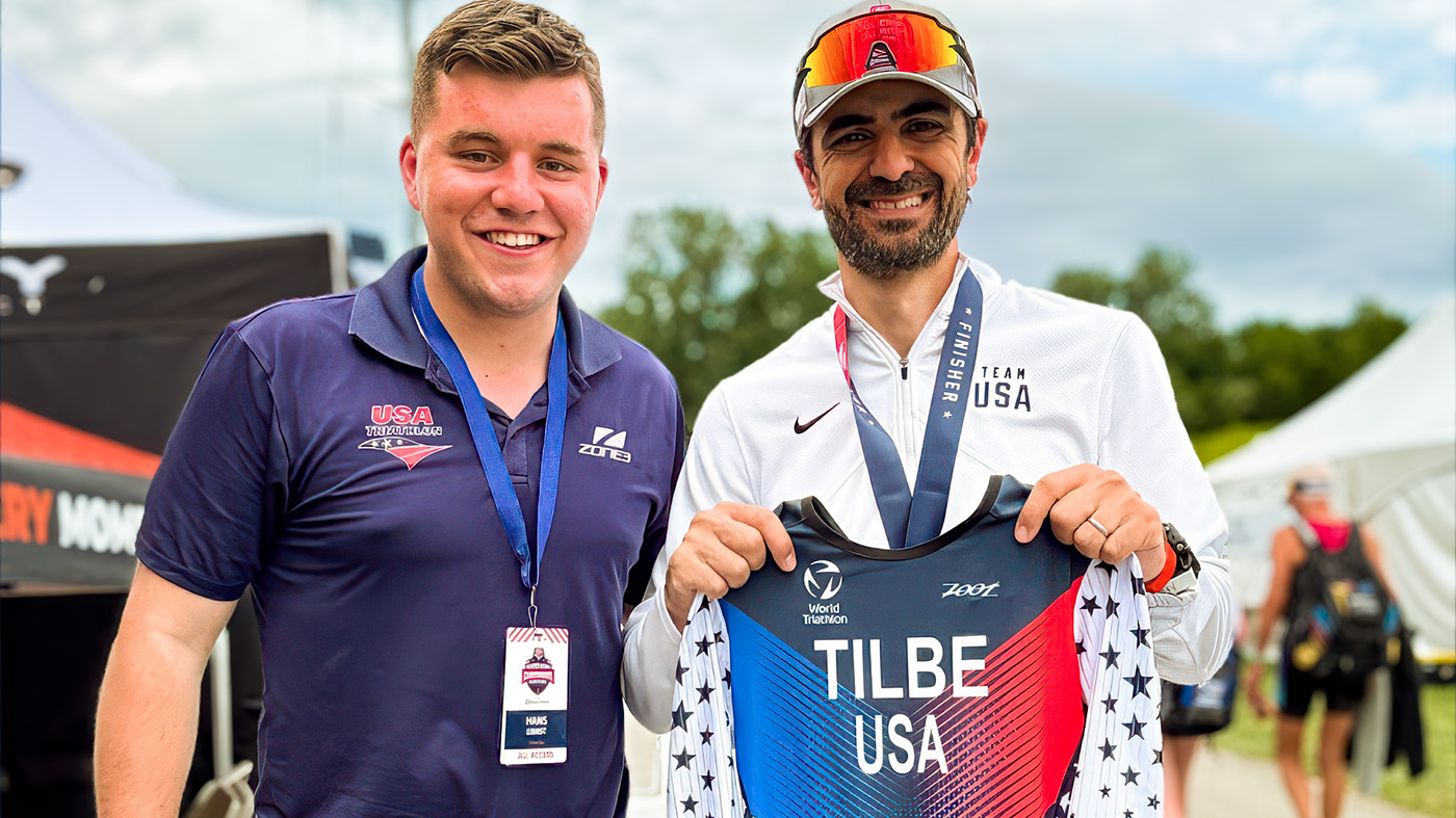 Anil Tilbe (right), smiles as he stands stands next to Team USA coordinator for Team USA Triathlon, Hans Ernst, holding a “Tilbe USA” athletic uniform.